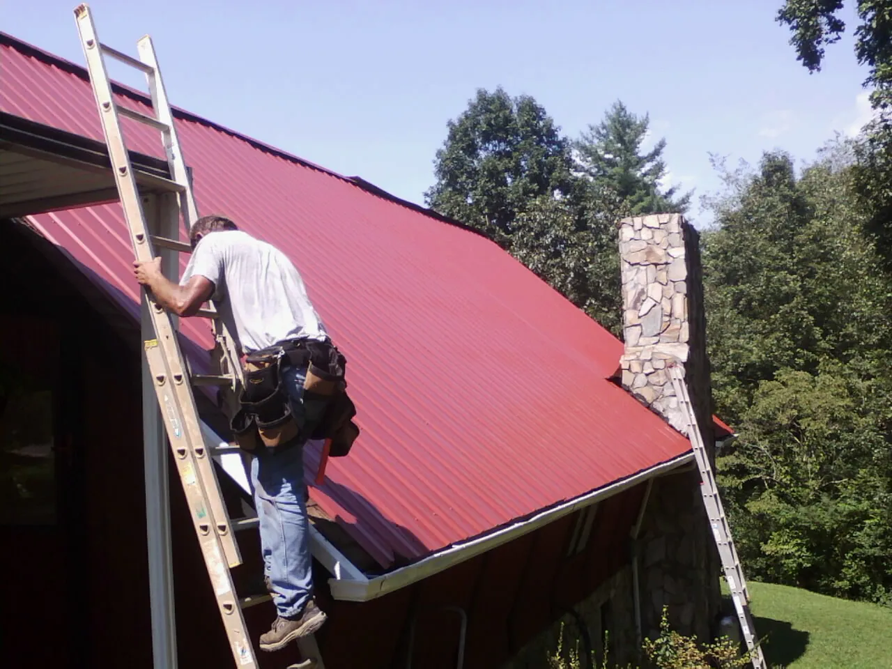 Roofer on a ladder beside a red metal roof and stone chimney — Soffit & Fascia Repair by Roofing Brothers in Crownsville